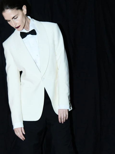 Elegant woman poses in a white tuxedo jacket, black bow tie, and black pants against a stark black backdrop, creating a striking contrast.