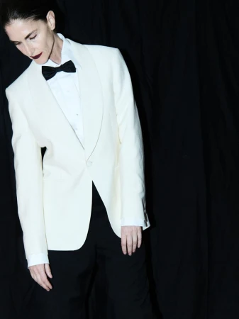 Elegant woman poses in a white tuxedo jacket, black bow tie, and black pants against a stark black backdrop, creating a striking contrast.