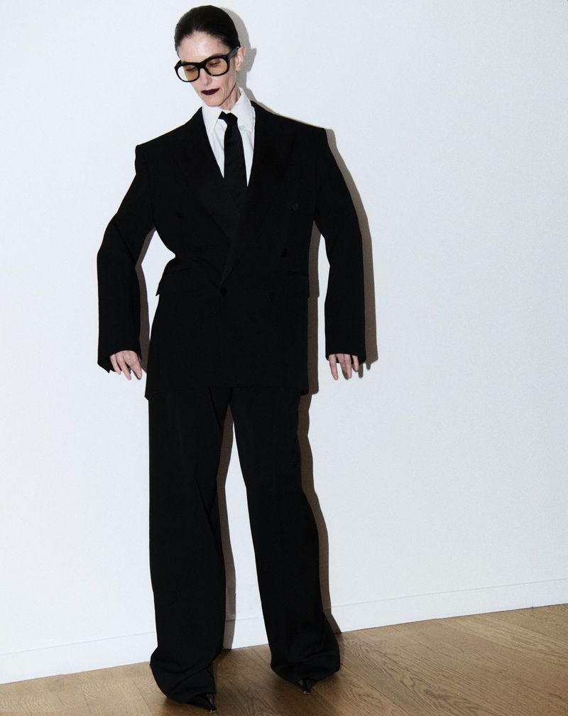 An adult woman stands in a photo studio wearing a black suit, black pointed shoes, and dark lipstick, against a white background.