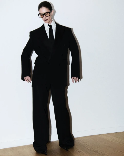 An adult woman stands in a photo studio wearing a black suit, black pointed shoes, and dark lipstick, against a white background.