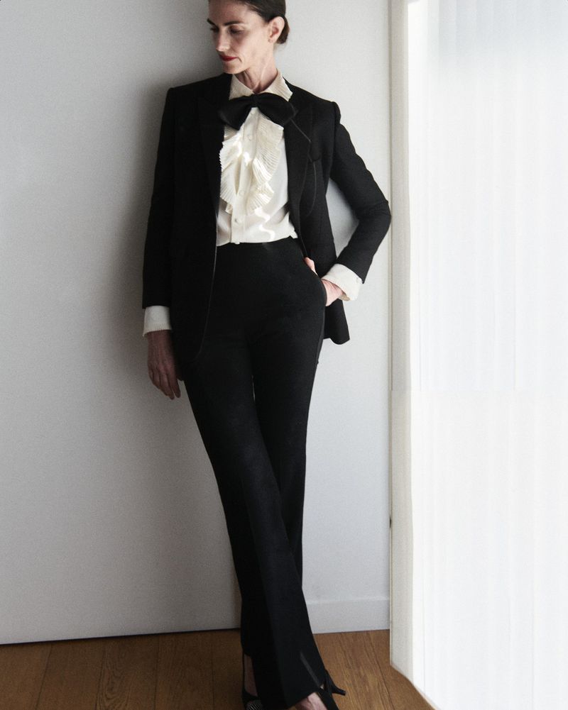 Elegant adult female in black suit with bow tie and ruffled blouse posing against a white wall indoors, fashion editorial-style portrait.