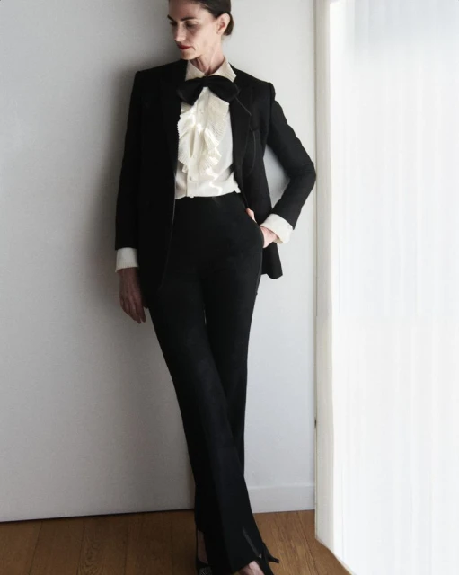 Elegant adult female in black suit with bow tie and ruffled blouse posing against a white wall indoors, fashion editorial-style portrait.