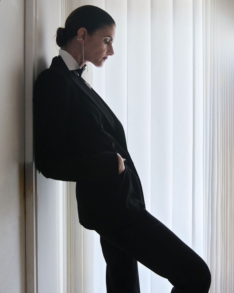 Elegant woman in a black tuxedo poses indoors near a window with vertical blinds, showcasing a blend of sophistication and modern fashion.