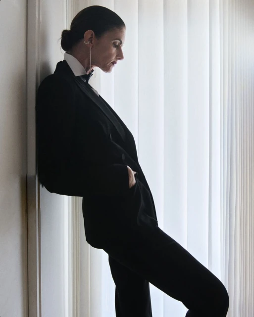 Elegant woman in a black tuxedo poses indoors near a window with vertical blinds, showcasing a blend of sophistication and modern fashion.