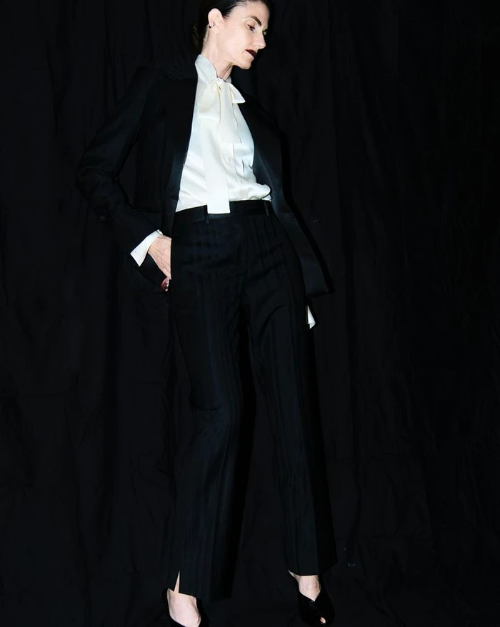 Woman in a black suit and white silk bow blouse standing against a dark fabric background for M Le magazine du Monde