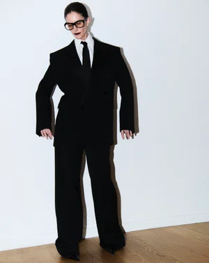 Model in black suit and tie wearing thick glasses against a plain background
