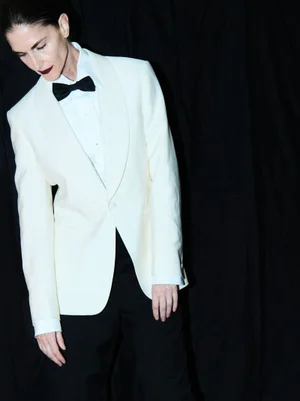 Model wearing a white tuxedo jacket, black bowtie, and dark trousers against a plain black backdrop.