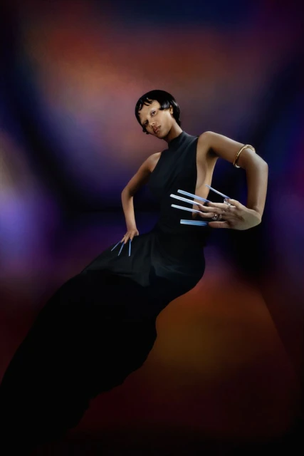 Woman in black dress with long nails posing against a colorful background, showcasing fashion and artistic expression in a studio setting.