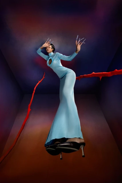 A woman with dramatic makeup and a light-blue dress poses in a room with a surreal and slightly ominous atmosphere, with high heels.
