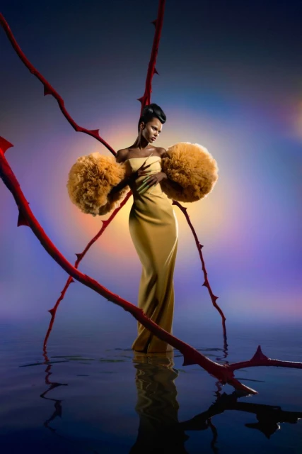 Elegant African-American woman in gold gown with exaggerated sleeves posing in water with red branches, vibrant background, fashion photography.