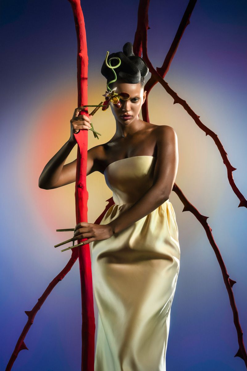 Fashion shot of a woman in a light-yellow dress with sculptural hair, holding a rose stem, with red branches in the background.