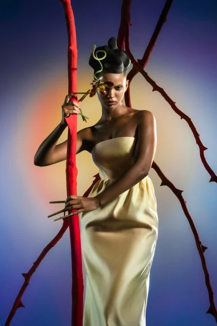 Fashion shot of a woman in a light-yellow dress with sculptural hair, holding a rose stem, with red branches in the background.