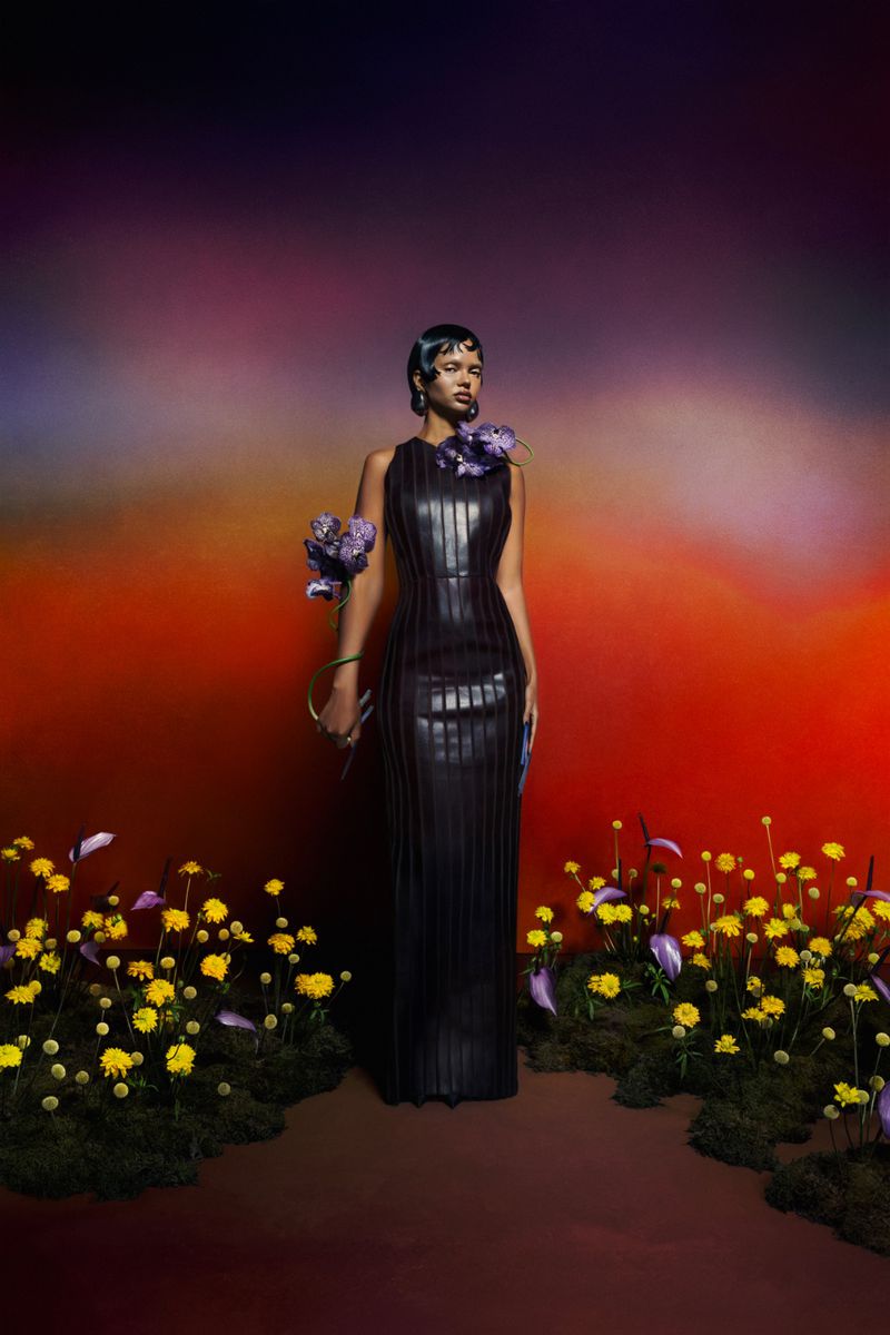 A woman wearing a black sleeveless gown with floral accents stands amidst yellow wildflowers against a gradient, colorful background.