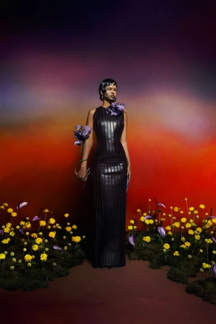 A woman wearing a black sleeveless gown with floral accents stands amidst yellow wildflowers against a gradient, colorful background.