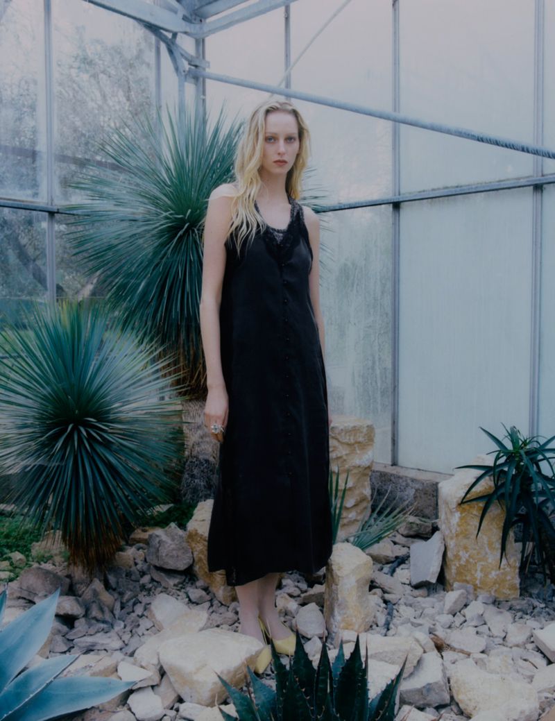 A tall, slender blonde woman in a black dress stands among desert plants and rocks in a greenhouse setting, showcasing a fashion editorial look.