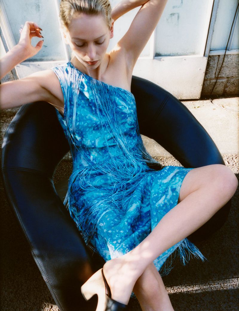 Woman in a blue one-shoulder fringe dress sitting in a black circular chair, wearing black heeled shoes and posing in a natural setting.