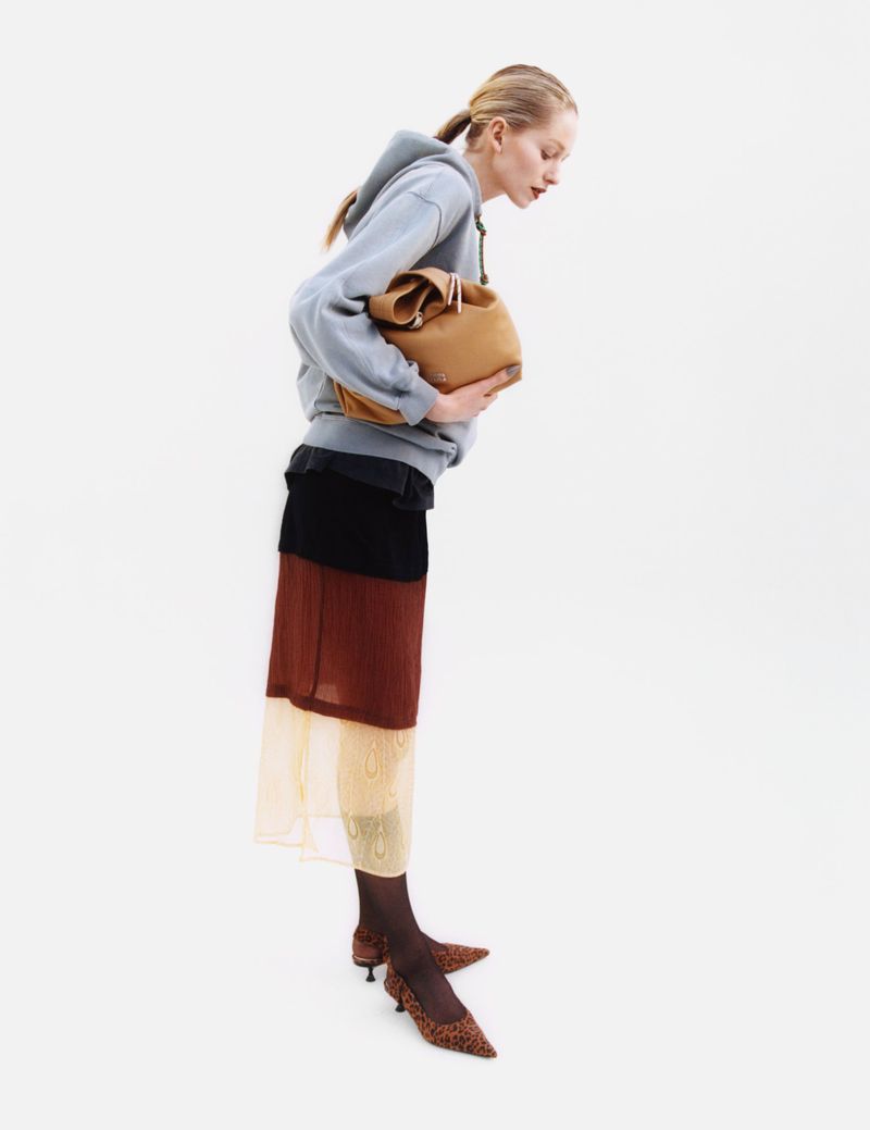 A woman in a gray hoodie and layered skirt holds a tan leather bucket bag in a studio shot against a bright white backdrop.