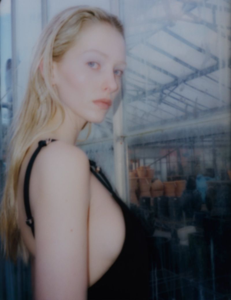 Close-up of a blonde woman in a black top looking over her shoulder, with a greenhouse-like structure visible in the background.