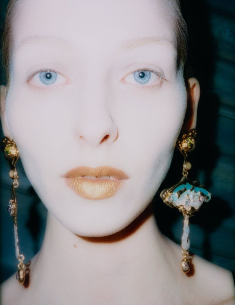 Close-up portrait of a woman with golden lips wearing ornate turquoise and gold earrings against a dark blue background.