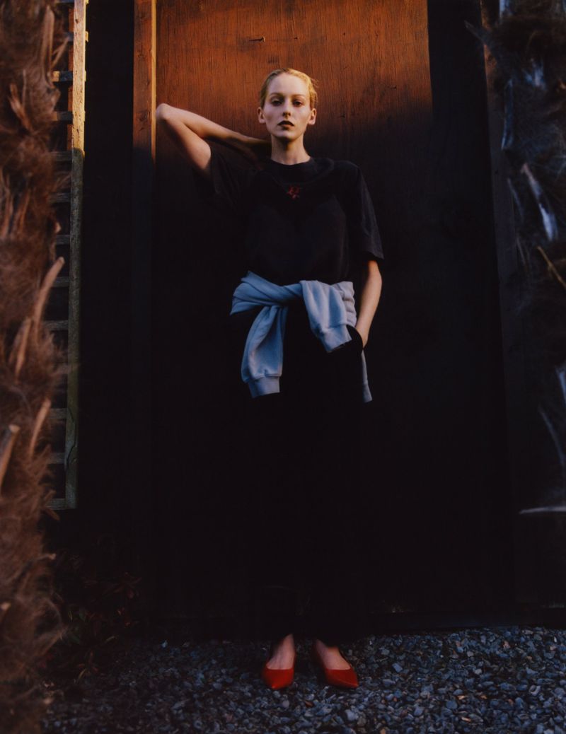 An adult woman poses against a dark door wearing a T-shirt, skirt, and red flats, with a light blue sweater tied around her waist outdoors.