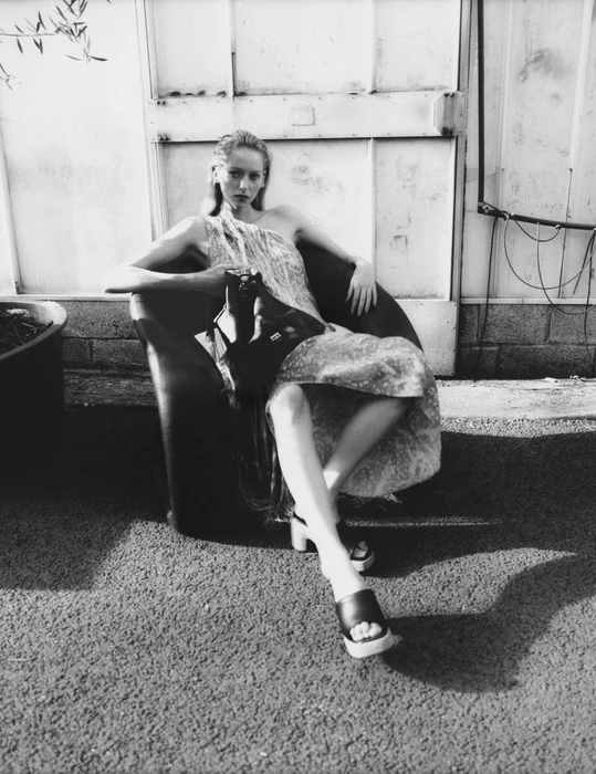 Model seated in a dark armchair wearing a patterned dress and holding a handbag in an outdoor setting