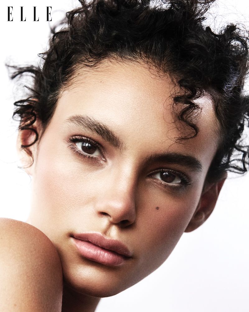 Close-up shot of a woman with curly hair and light makeup on a white background. Elle Magazine logo visible in the upper left.