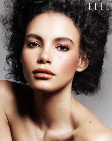 Close-up portrait of a woman with dark curly hair and light skin, wearing makeup, set against a neutral grey backdrop. Elle logo visible.