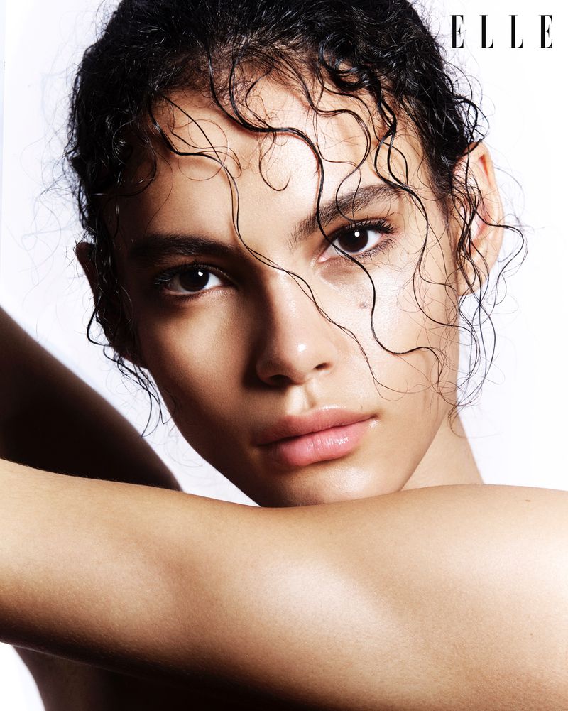 Close-up portrait of a young adult woman with dark curly wet hair, arms crossed near her face, featuring natural makeup and an Elle magazine logo.