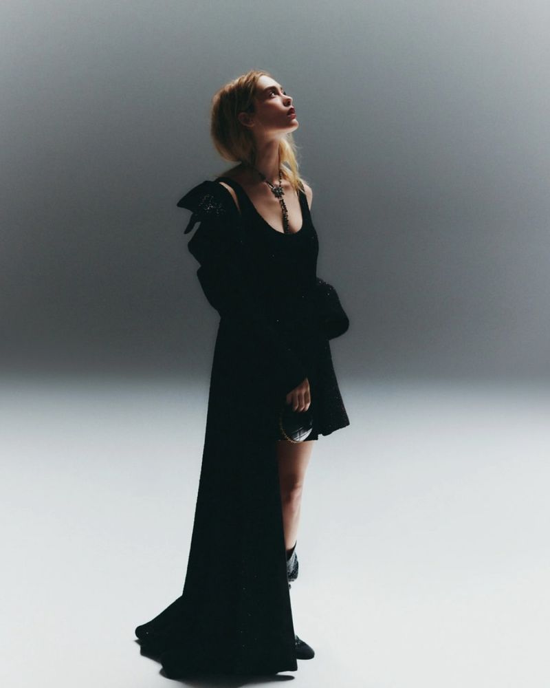 A young fair-skinned woman in a dark dress and boots poses in a brightly lit photo studio, looking upward with a slight smile.