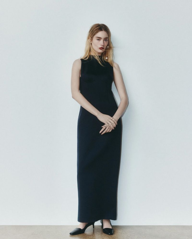 Elegant woman in a black sleeveless column dress stands against a white backdrop, complemented by black heeled shoes.