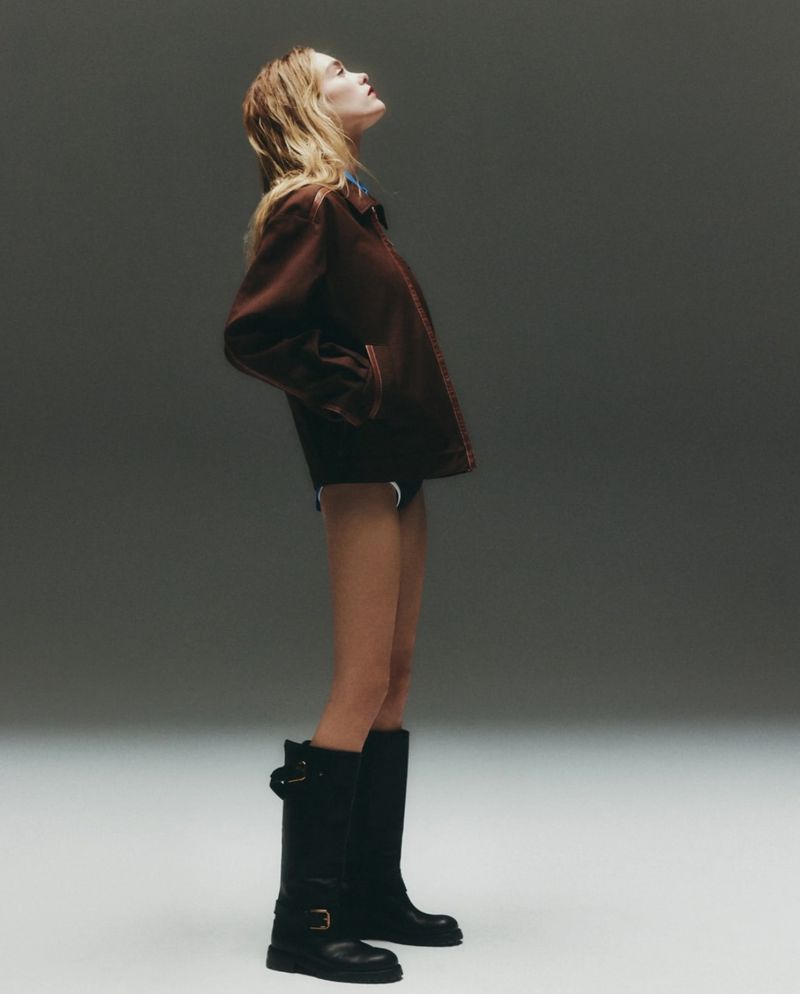 A woman with long blonde hair poses in a studio wearing a brown jacket, short bottoms, and black boots, looking up and away from the camera.