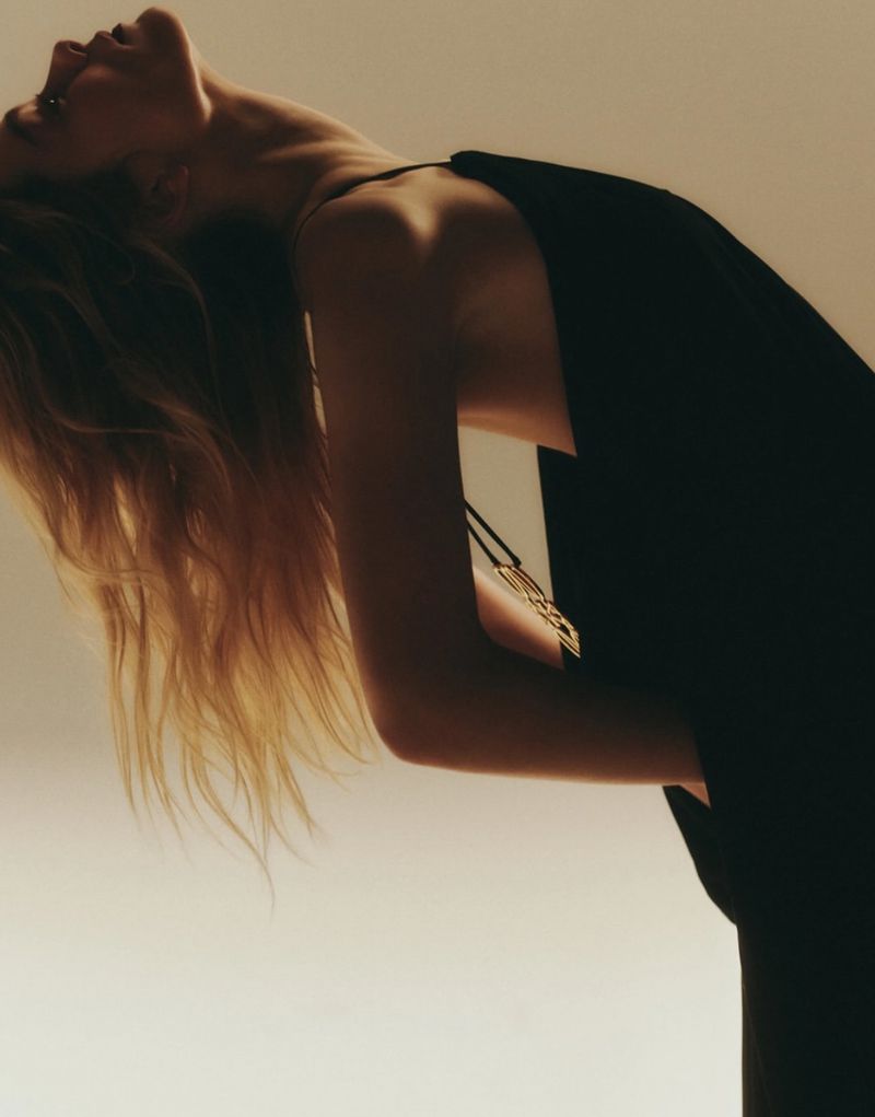 Woman in black dress with head tilted back shows off long blonde hair against a gradient background in an elegant studio shot.