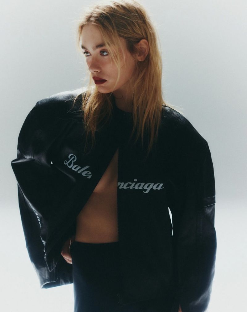 A blonde woman poses in a black Balenciaga jacket and top against a white background in a fashion editorial shot indoors.