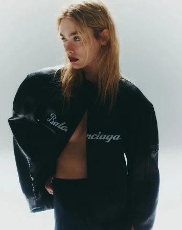 A blonde woman poses in a black Balenciaga jacket and top against a white background in a fashion editorial shot indoors.
