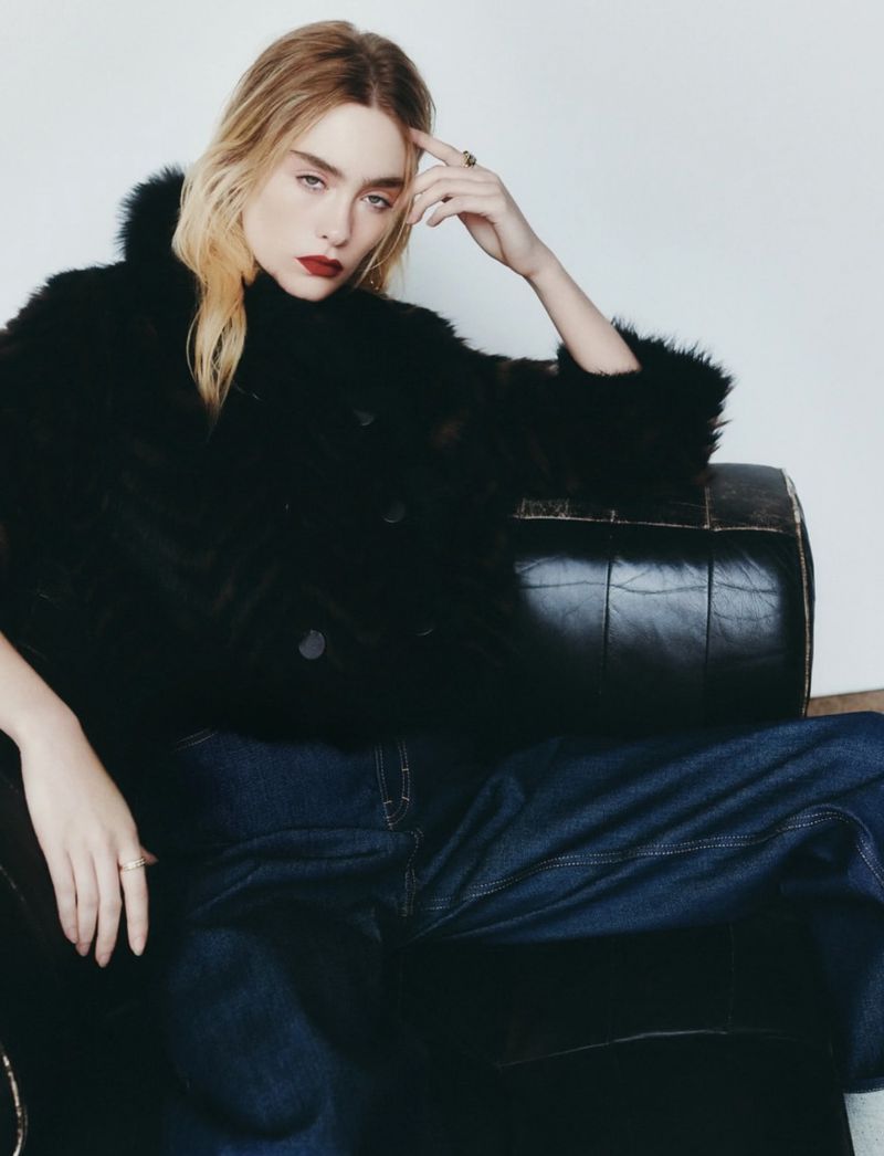 A fair-skinned adult woman with red lips poses in a black furry jacket and jeans, sitting in a black leather armchair indoors.