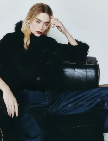 A fair-skinned adult woman with red lips poses in a black furry jacket and jeans, sitting in a black leather armchair indoors.