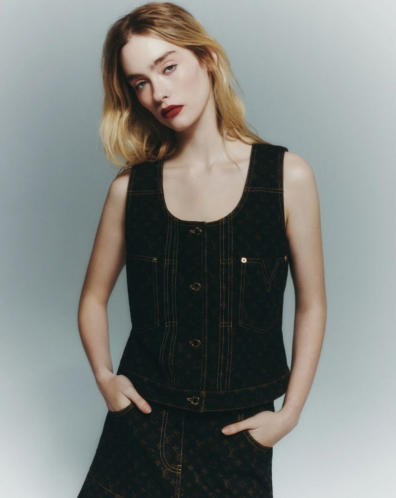 Woman in a dark patterned denim vest and skirt poses in a studio with fair skin, blonde hair and dark red lipstick with her hands in her pockets.