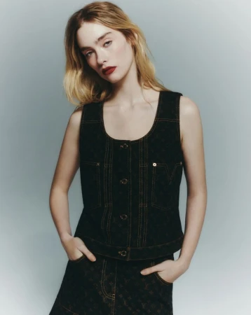 Woman in a dark patterned denim vest and skirt poses in a studio with fair skin, blonde hair and dark red lipstick with her hands in her pockets.