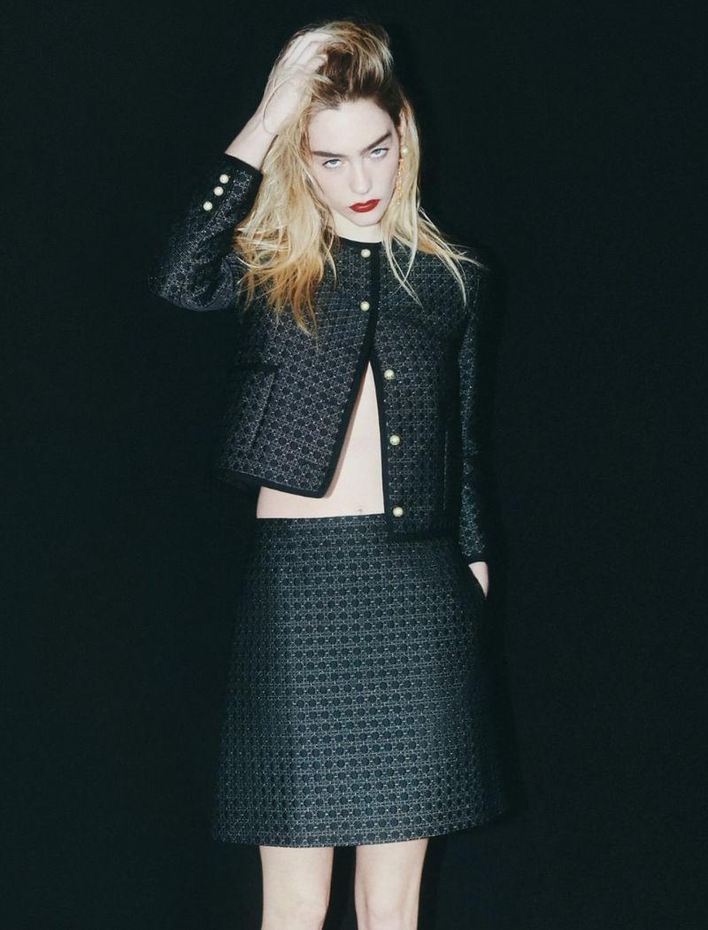 Fashionable young woman poses in a patterned black skirt and jacket against a dark backdrop, showcasing a modern, elegant style.