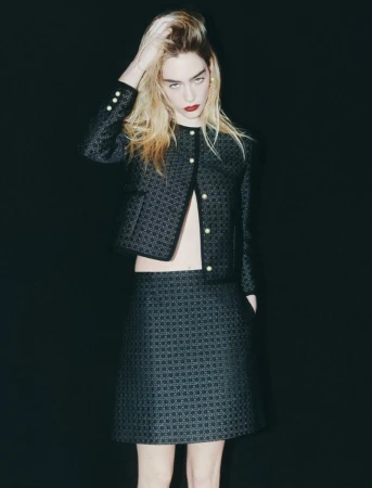 Fashionable young woman poses in a patterned black skirt and jacket against a dark backdrop, showcasing a modern, elegant style.