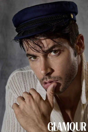 Portrait of a male model with light brown hair, wearing a blue sailor cap and a loosely buttoned white linen shirt in a photo studio.