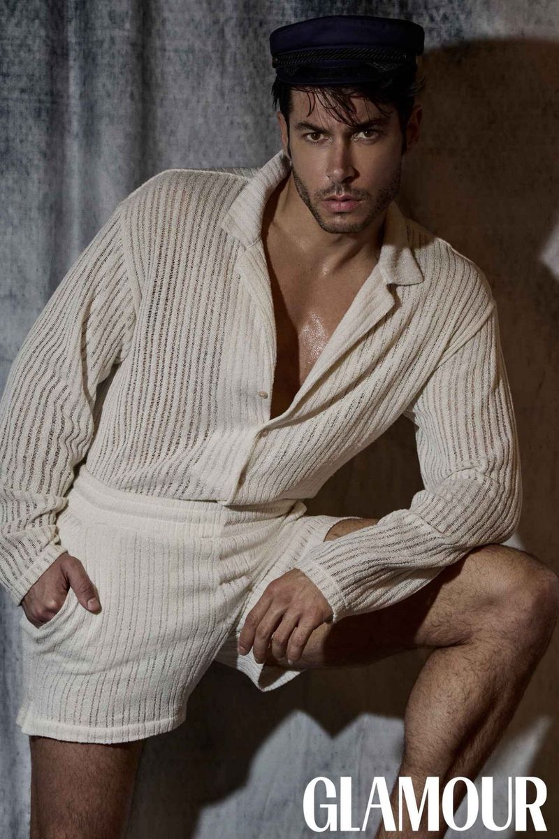 Portrait of a young adult male in a relaxed pose, wearing a short and shirt, accessorized with a dark cap, set against a textured background.