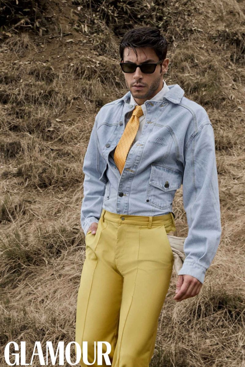 An adult man in a blue jacket and yellow pants stands in a field, wearing sunglasses and a tie, posing for a fashion editorial.
