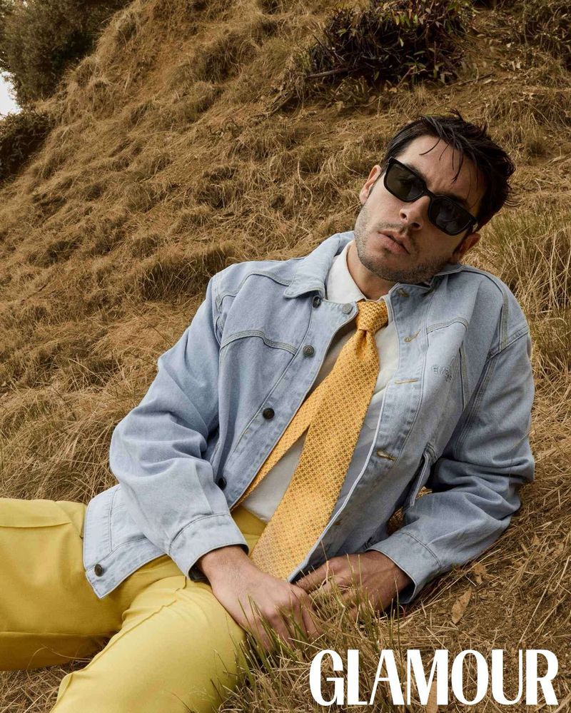 Man in yellow pants and tie with a blue jean jacket reclining in dry field with the Glamour magazine logo at the bottom.