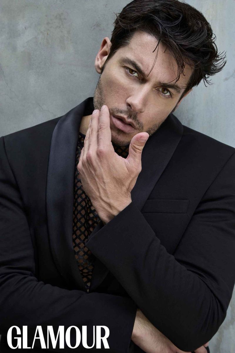 Portrait of a man in a black suit and mesh top with hand to his face in front of gray studio backdrop; Glamour logo visible.