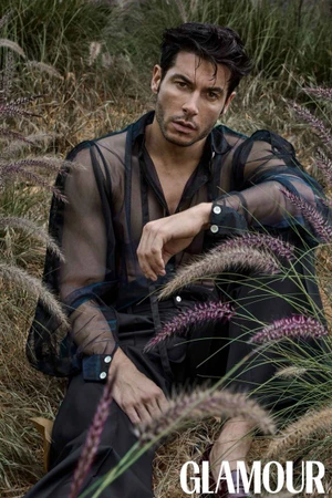Model Andrea Denver posing amidst tall grasses in a sheer black shirt for Glamour Bulgaria
