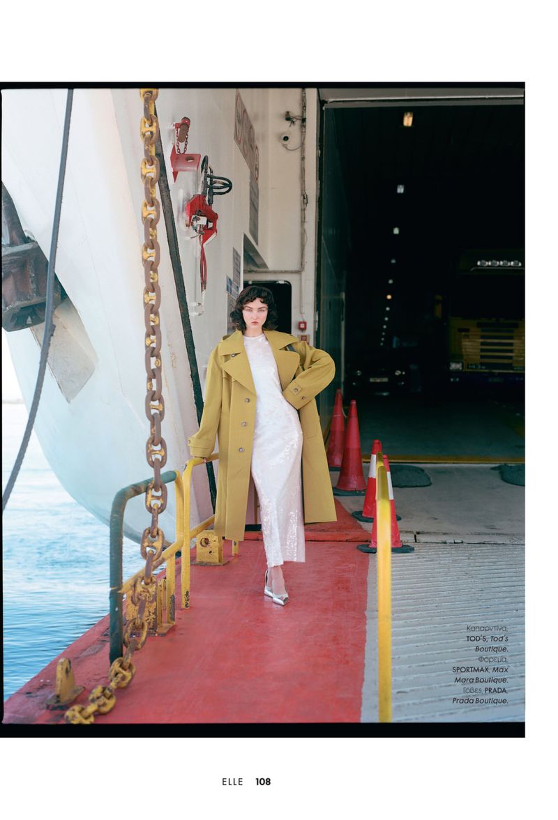 Fashionable woman in a white dress and yellow coat poses on a ferry with a neutral facial expression, exhibiting editorial style.