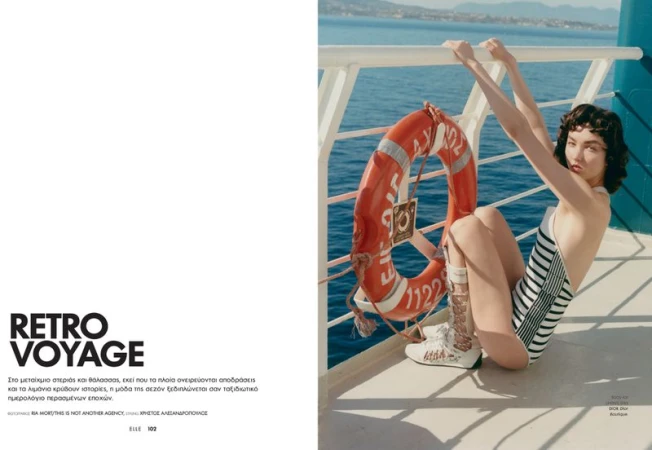 Woman in striped bathing suit poses next to orange life preserver on a boat with blue ocean and distant land in background, fashion editorial.
