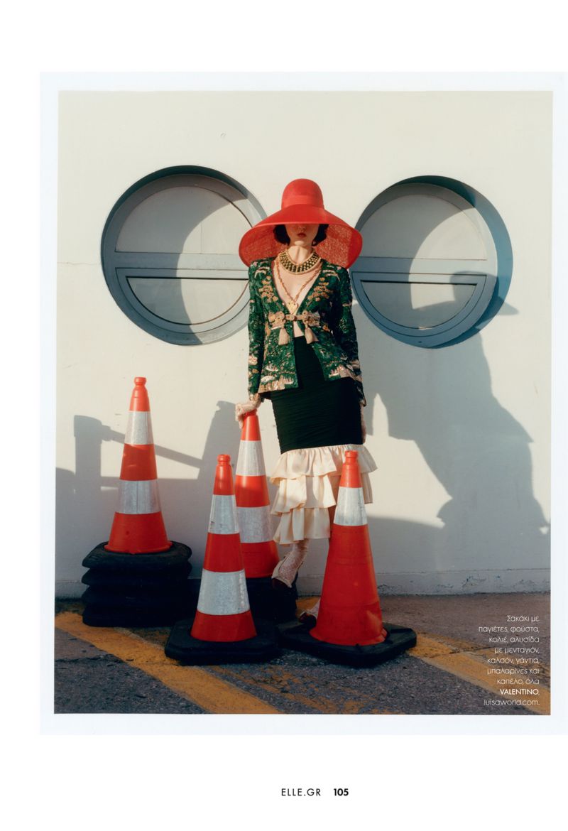Fashion editorial shot of a woman in oversized hat and green jacket posing with orange traffic cones on a city street.