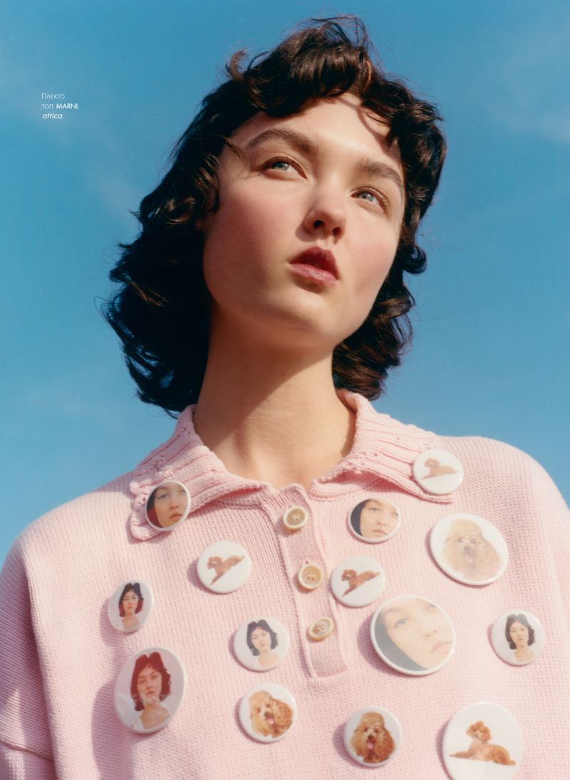 Low-angle shot of a brunette woman wearing a pink collared shirt decorated with badges against a vibrant blue sky backdrop.